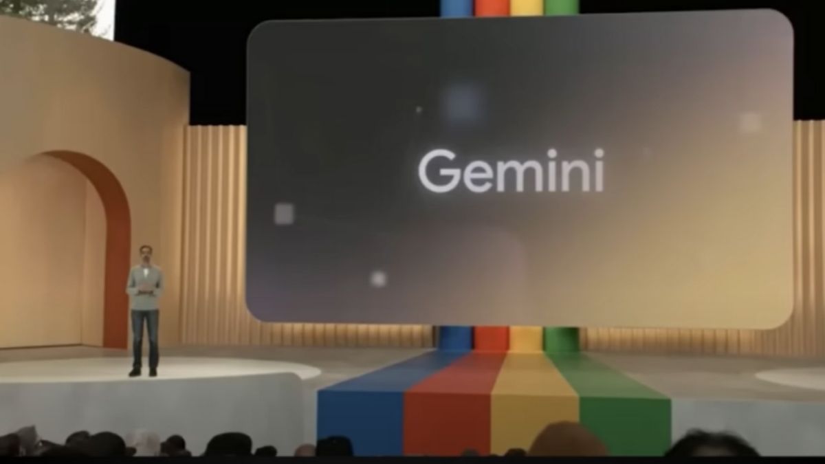  A man stands on a stage in front of a large screen with the word 'Gemini' on it.